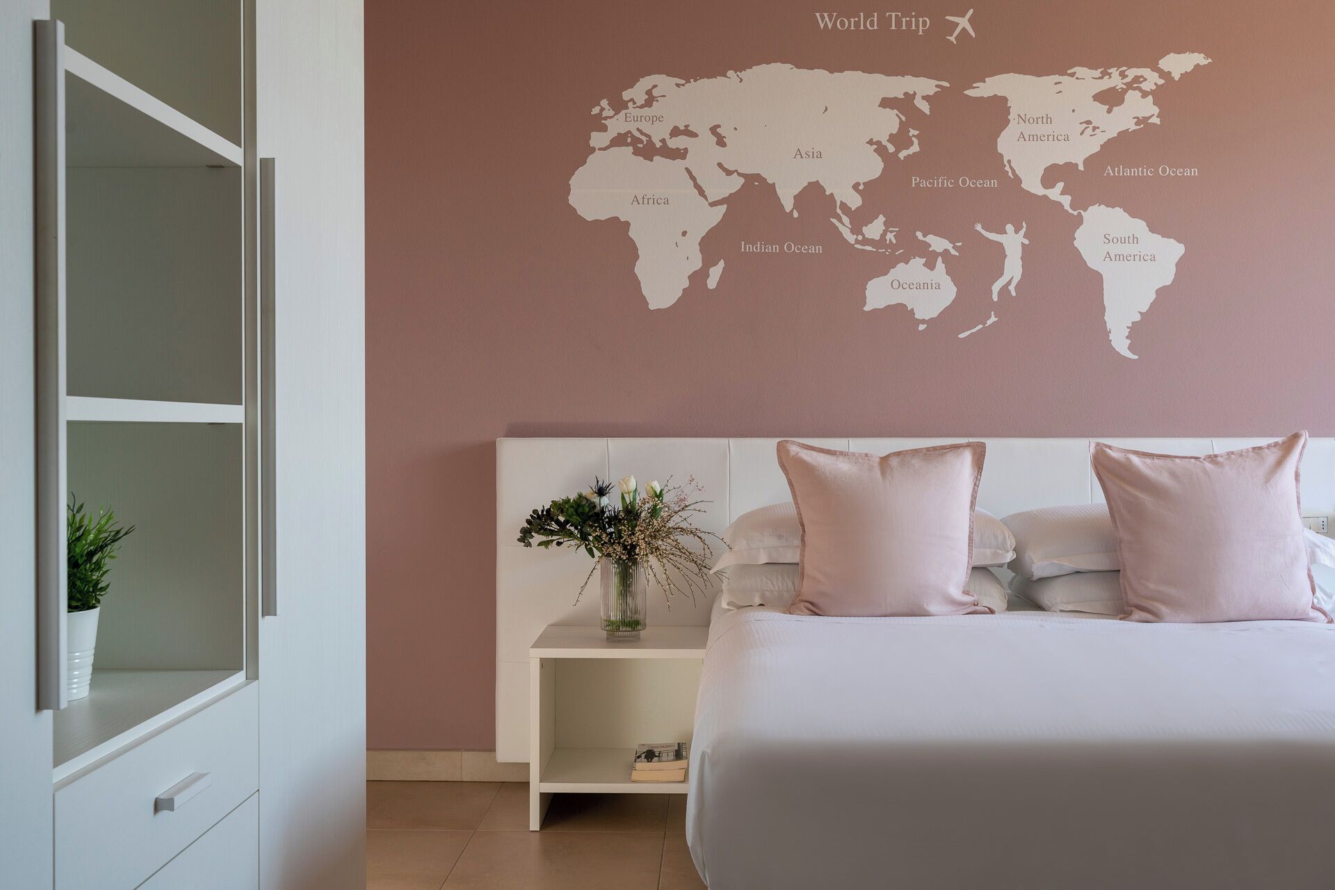 Een slaapkamer met een wereldkaart aan de muur, een bed met kussens, een nachtkastje met een bloevas en een witte kast.