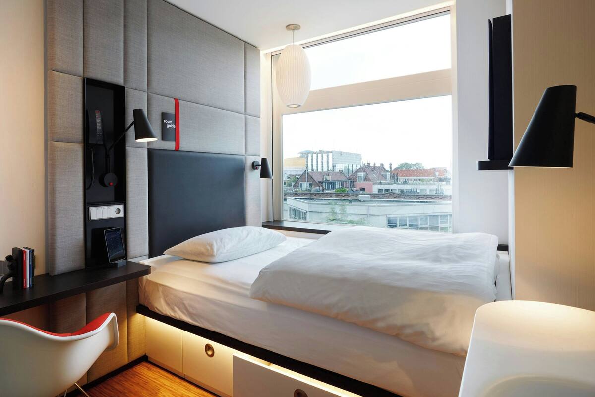 A modern hotel room with a bed, desk, and chair, a large window with a city view, and a mounted TV.