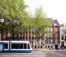 A tram passing by a row of traditional brick buildings with large windows and a tree in the foreground.