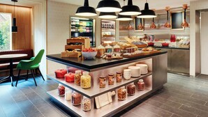 A breakfast bar with a variety of food items and condiments, a seating area with a green chair, and a kitchen area with hanging lights. - citizenM Amstel Amsterdam (Amsterdam)