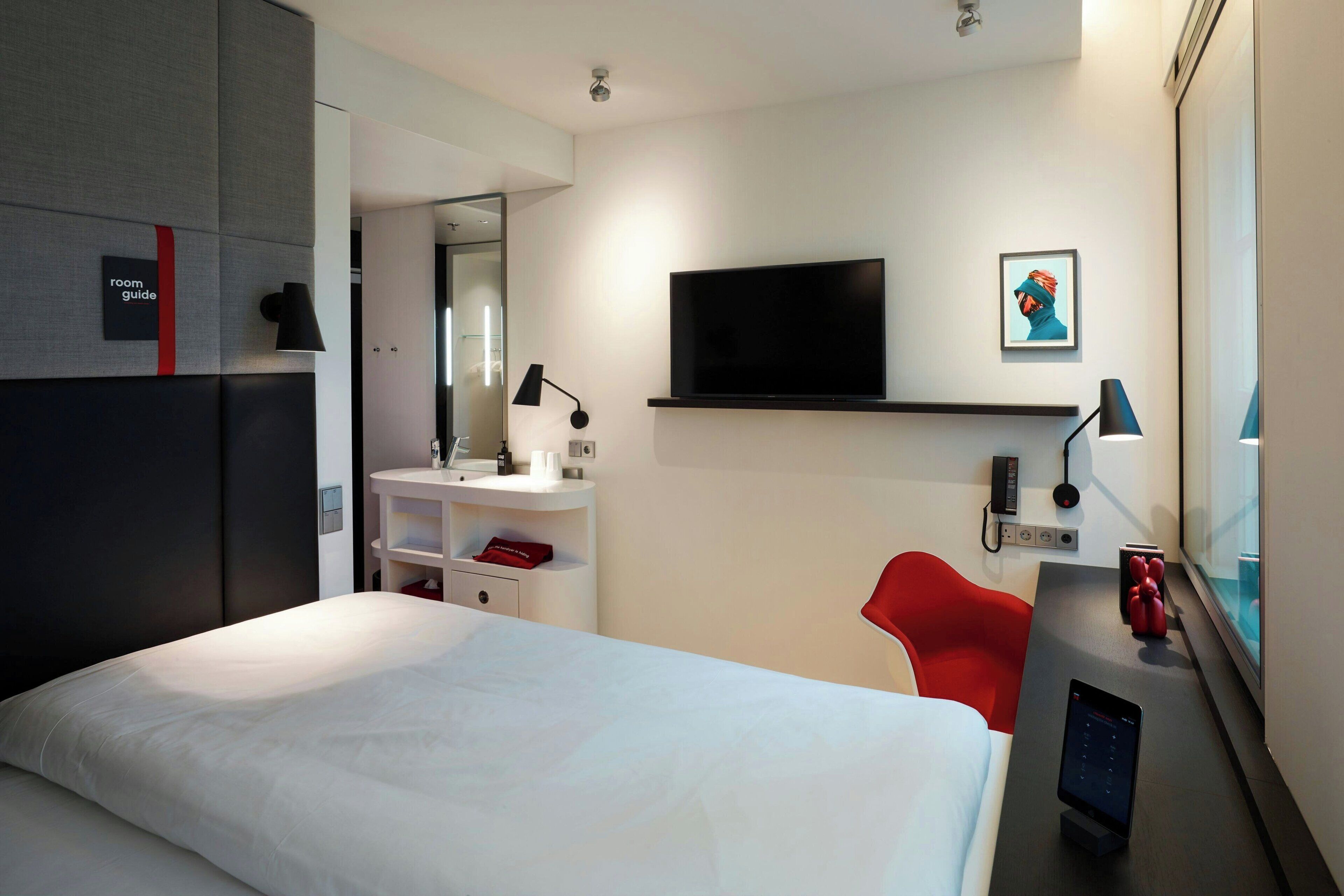 A hotel room with a bed, a desk with a computer, a TV, a red chair, and a bathroom with a mirror and sink.
