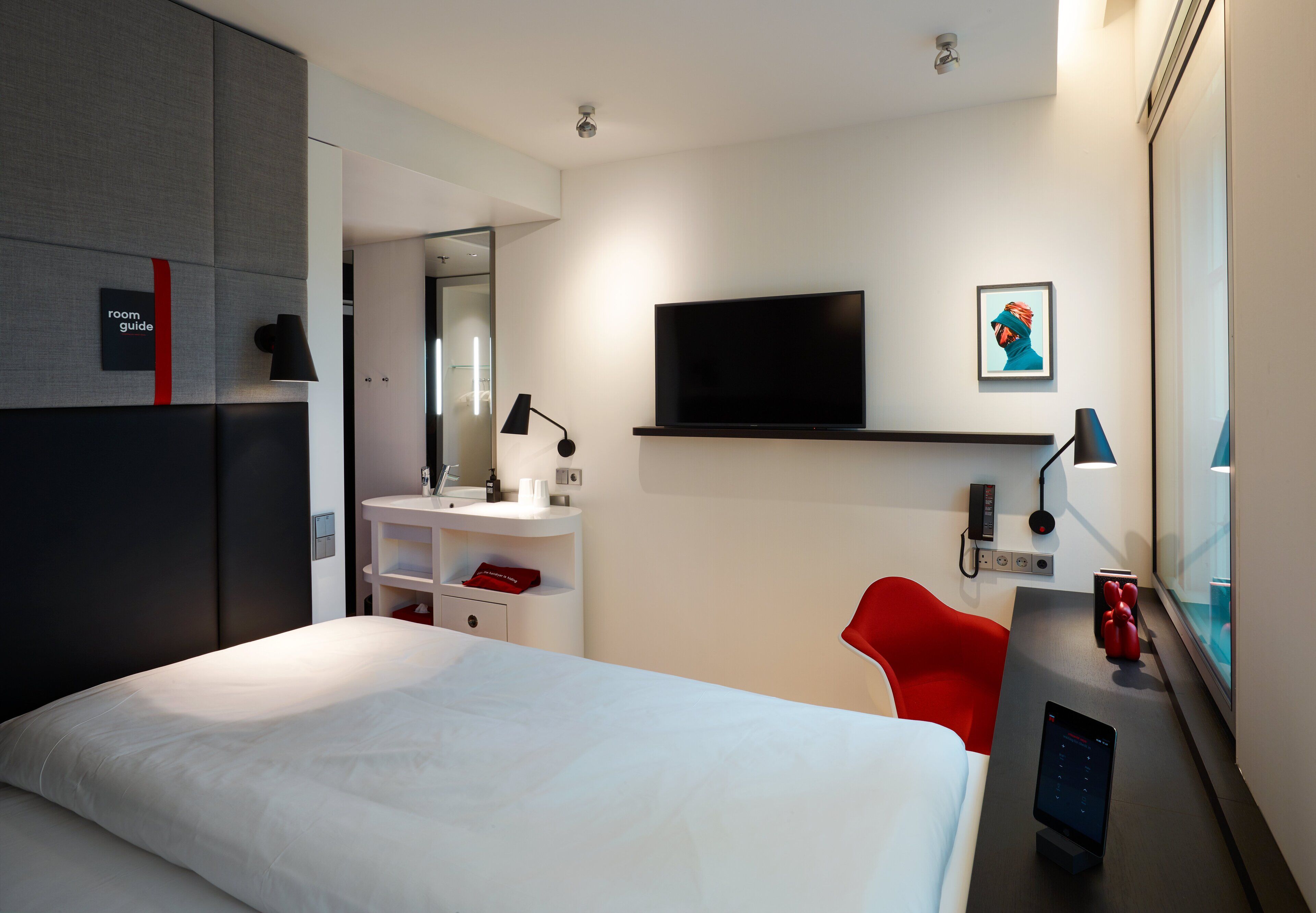 A modern hotel room with a large bed, a desk with a computer, a red chair, a TV, and a bathroom with a mirror and sink.