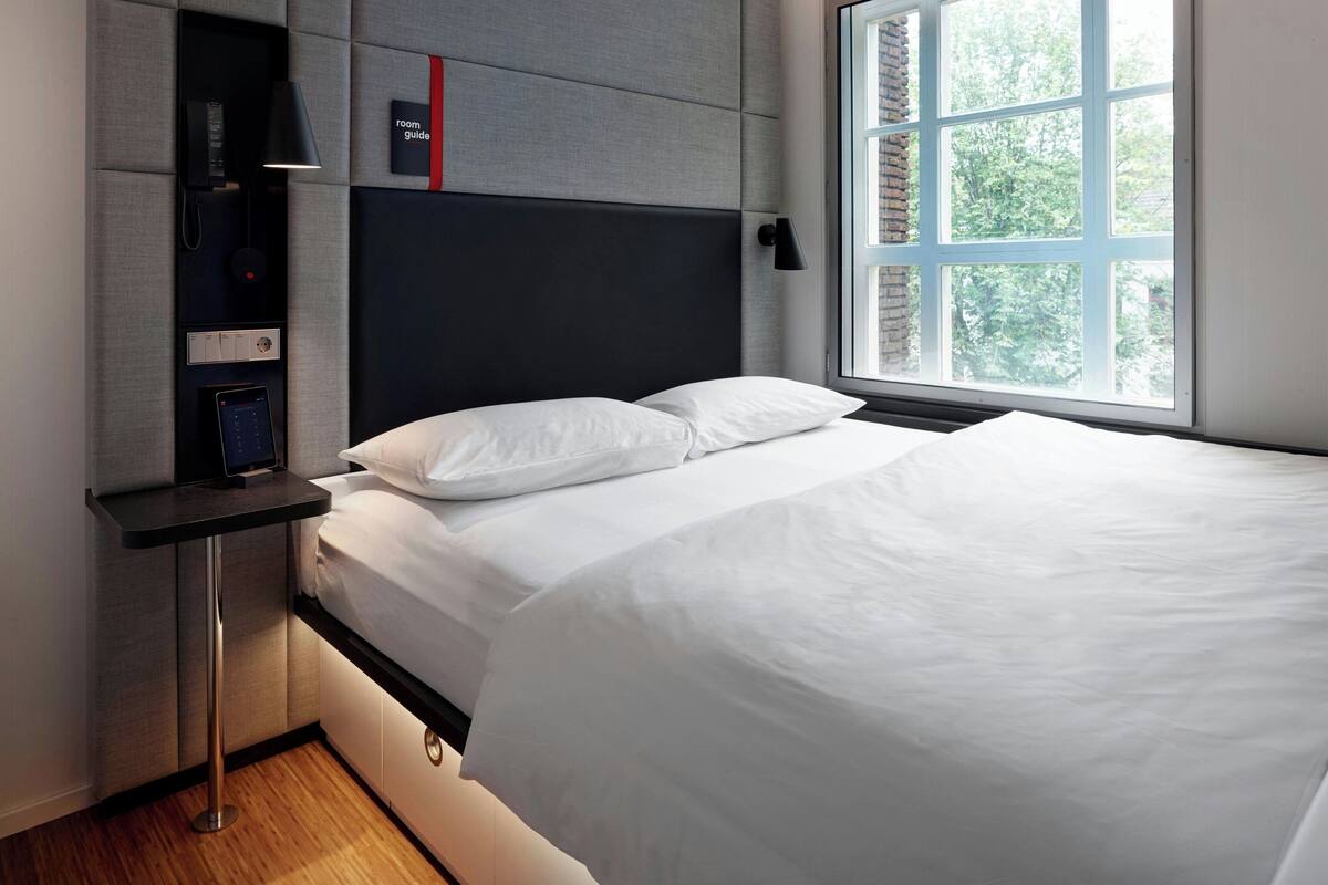 A modern hotel room with a bed, a bedside table, a built-in telephone, and a window with a view of trees.