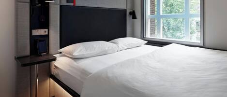 A modern hotel room with a bed, a bedside table, a built-in telephone, and a window with a view of trees.