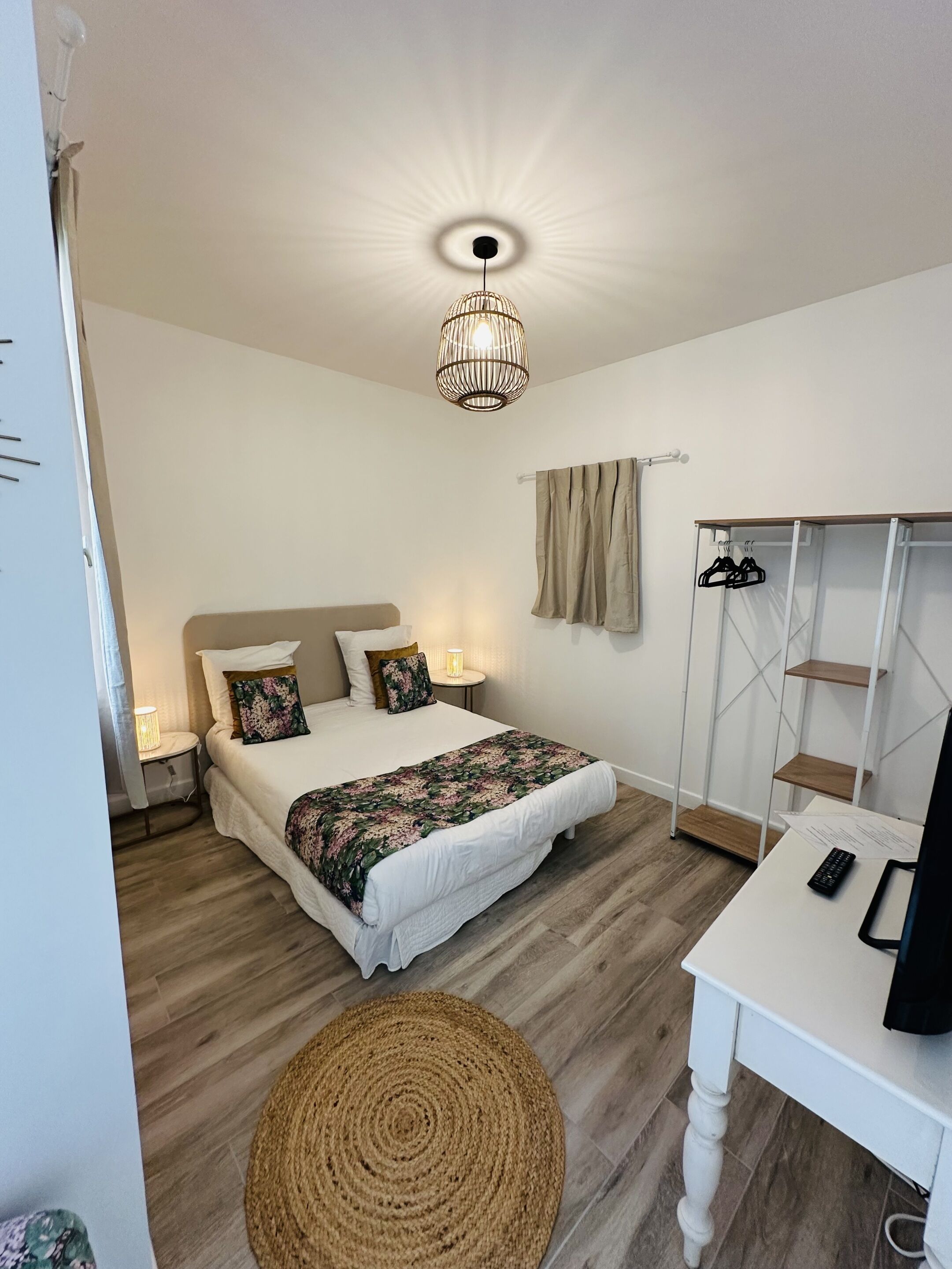 Un dormitorio con cama, mesitas de noche, escritorio, silla, alfombra tejida, armario y lámpara colgante.