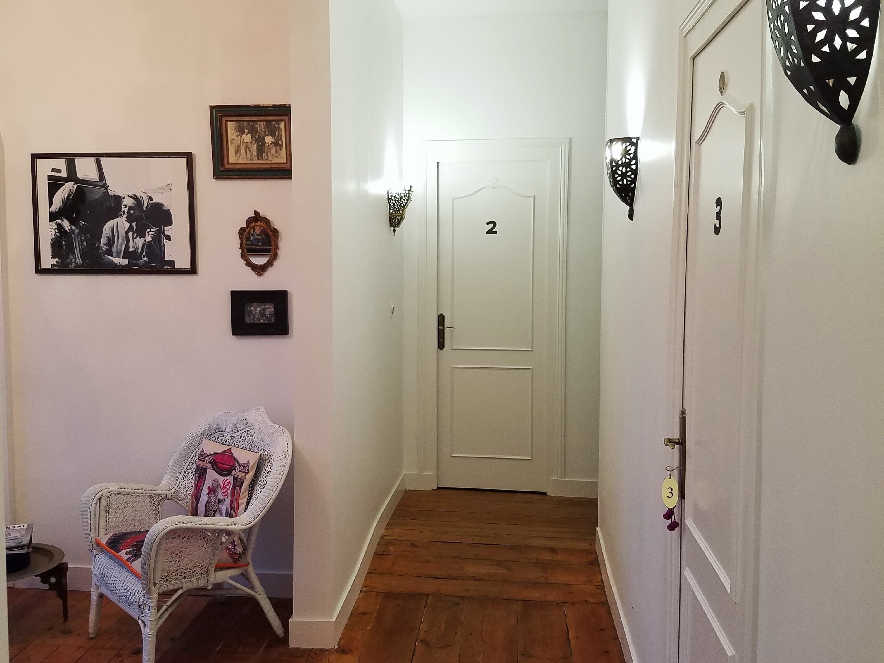 A hallway with a white door marked '2', a wicker chair with a cushion, and a wall adorned with framed pictures.