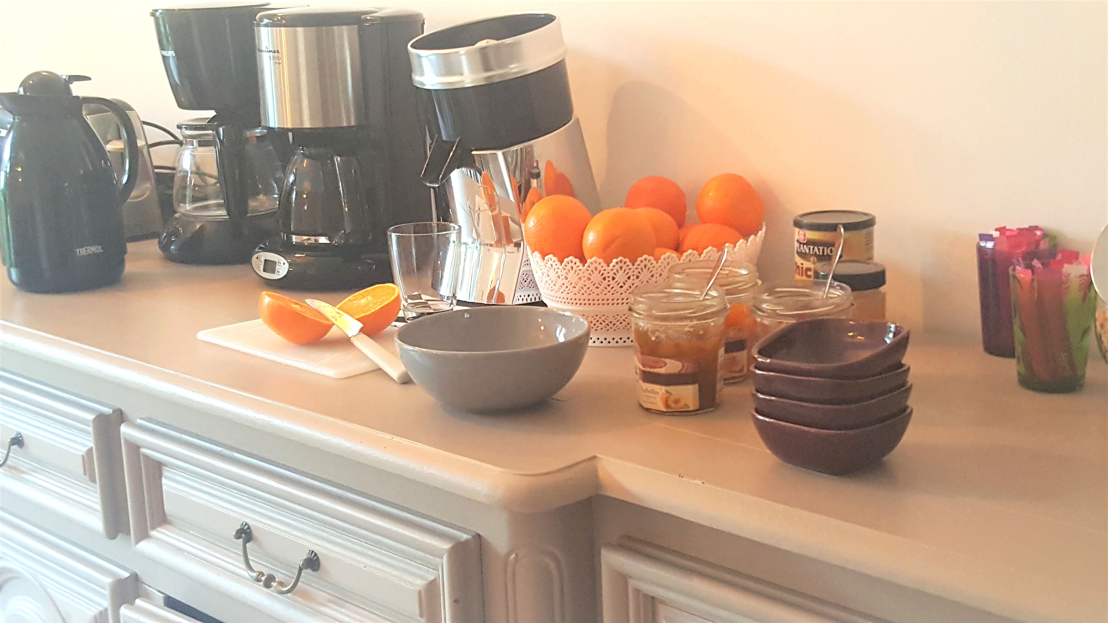 A kitchen counter with various appliances, a bowl, a basket of oranges, and jars.