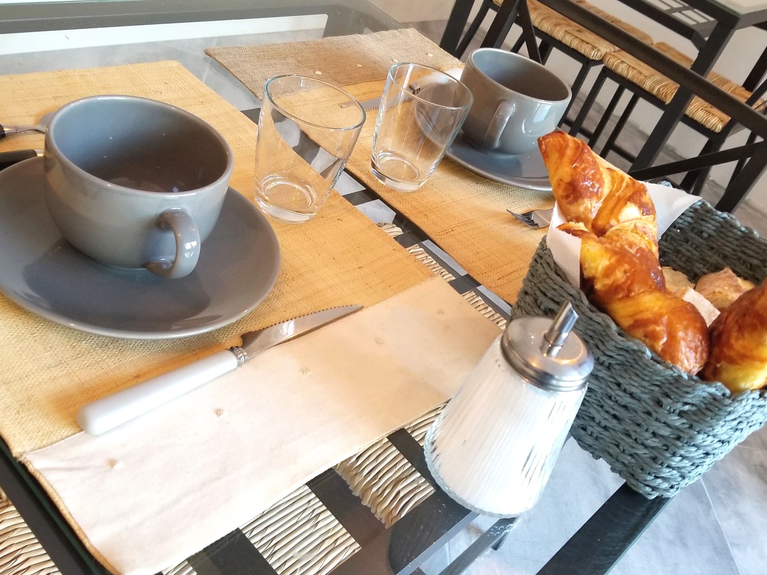 Estrat de taula per esmorzar, amb una tassa de cafè, dues copes, un ganivet, un salzet i un cisteller de croissants.