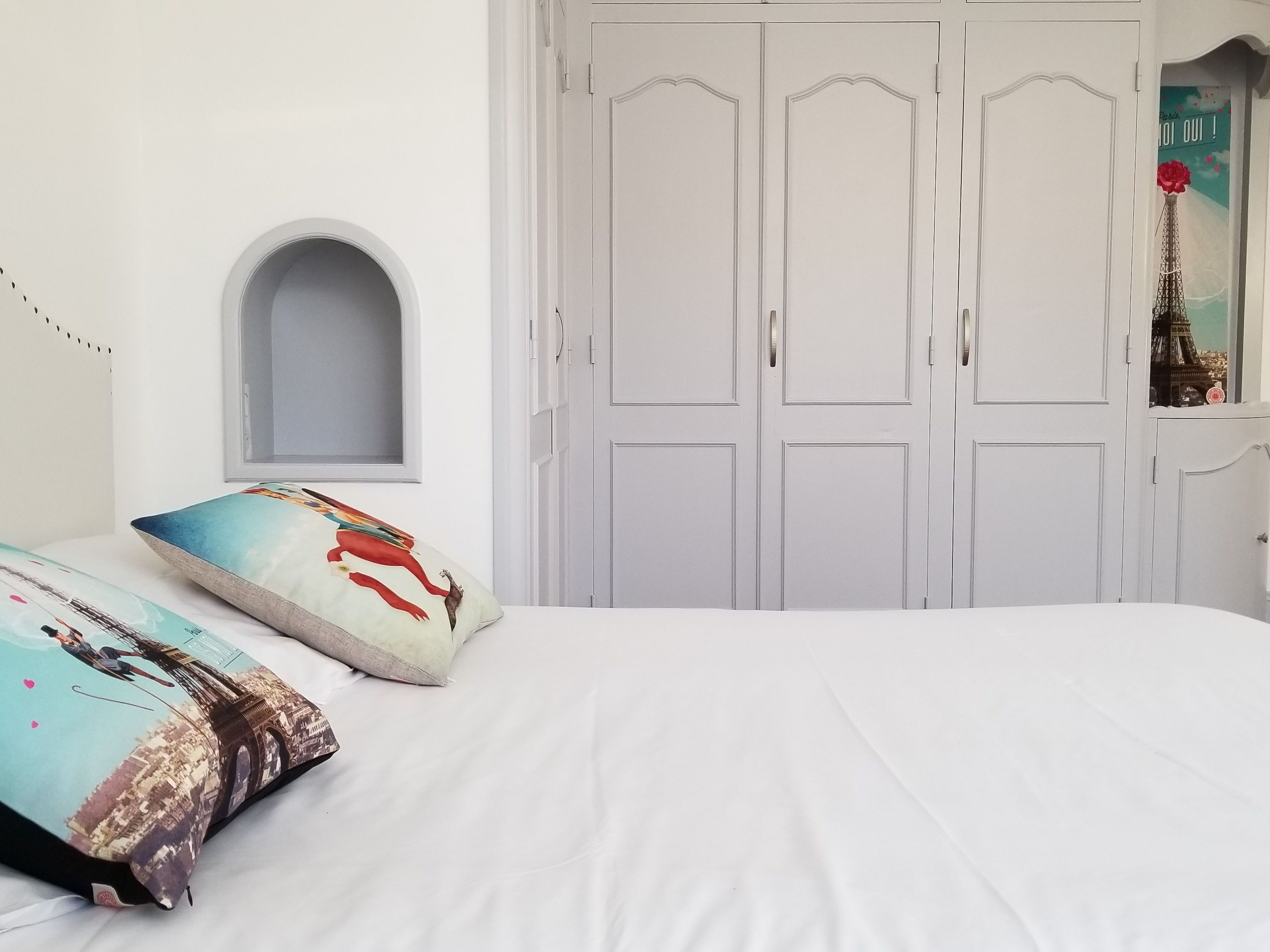 A bedroom with a bed, two pillows, a wardrobe, and a small arched window.