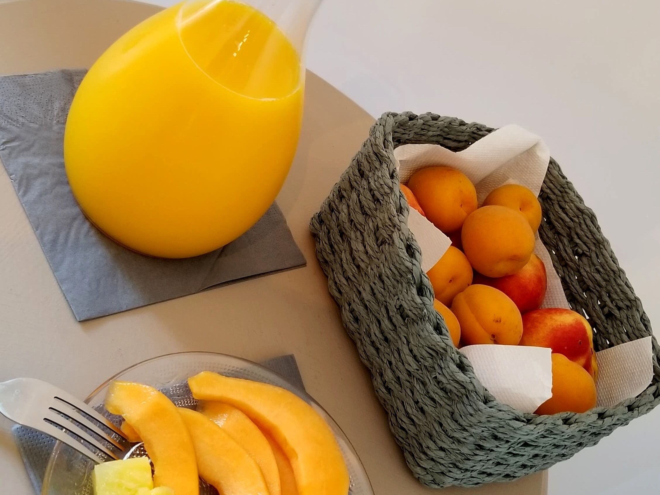 Una mesa con un vaso de jugo de naranja, una cesta de frutas y un plato con fruta cortada.