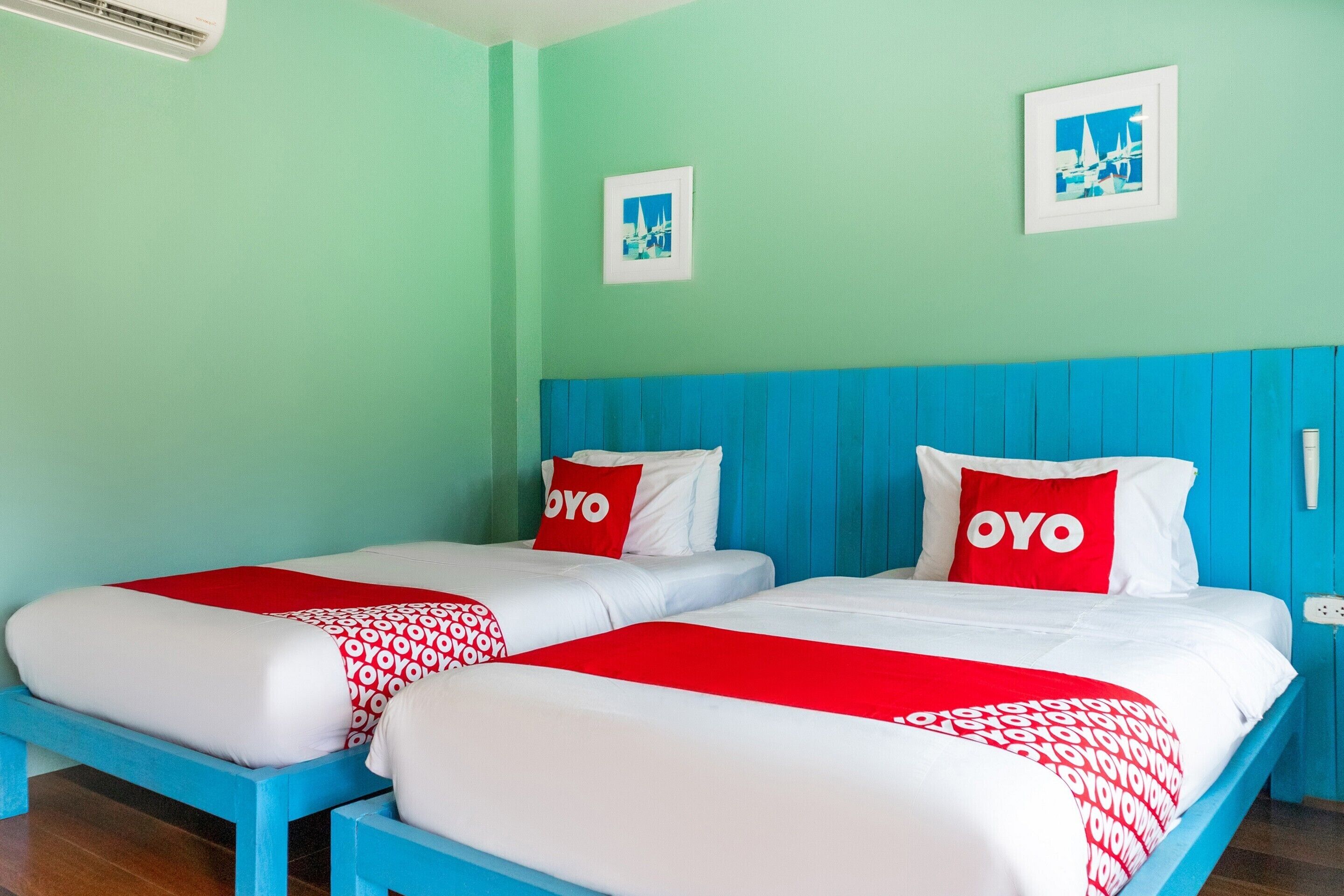 A room with two beds, each with a red and white bedspread and pillows with the word "OYO" on them. The room has light green walls and a blue headboard.