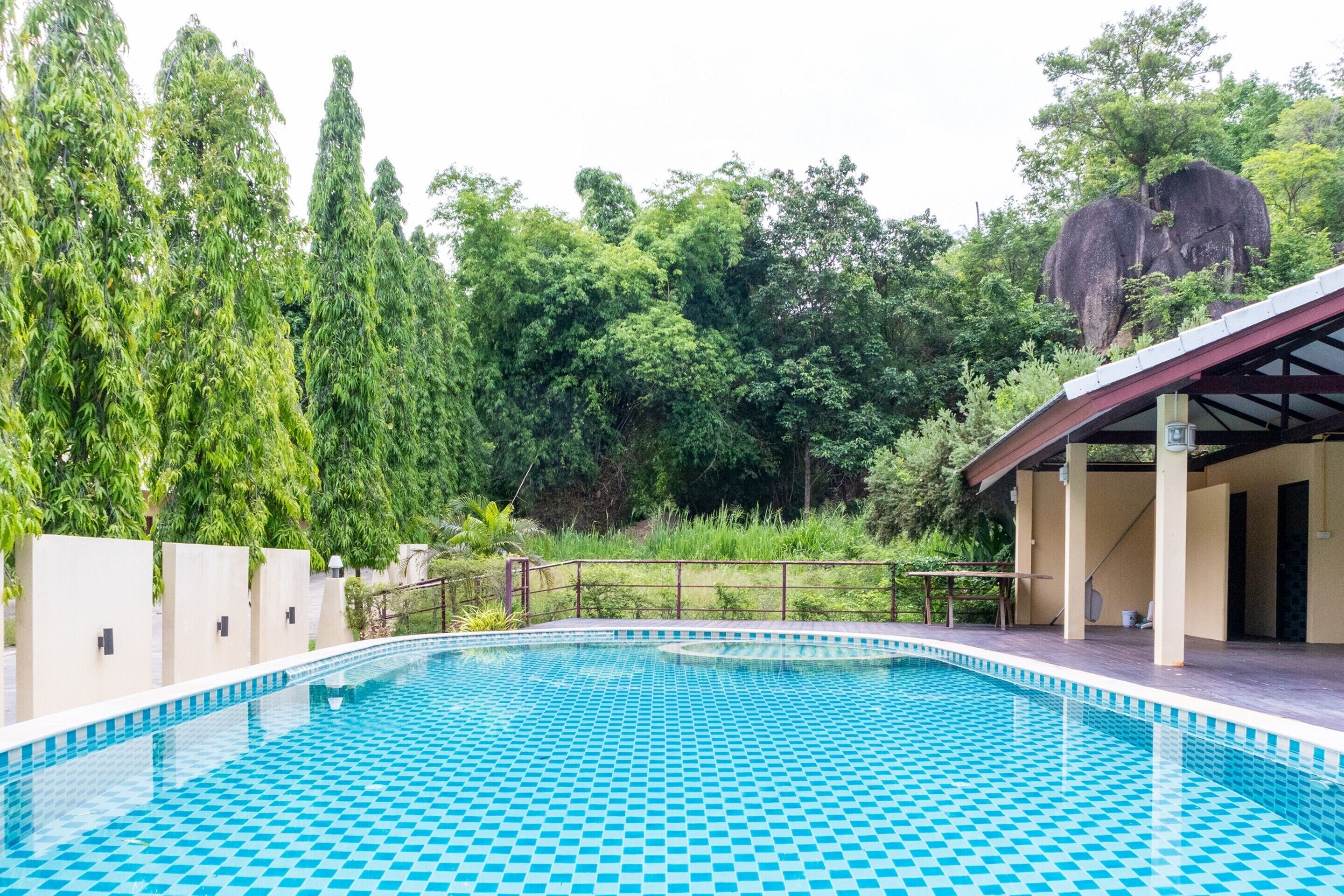 A swimming pool with a checkered pattern, surrounded by greenery and a covered area with pillars.