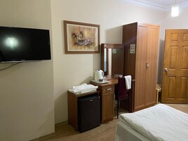 Habitación de hotel con cama, escritorio con computadora, refrigerador pequeño, una silla, televisor montado en la pared y armario de madera.