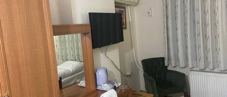 Une chambre d’hôtel avec un bureau en bois, un téléviseur fixé au mur, un fauteuil vert, un radiateur et une fenêtre avec des rideaux.