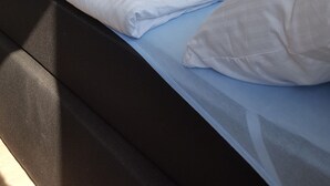 Ein ordentlich gemachtes Bett mit weißen Leinentüchern und zwei aufgerollten Handtüchern, ein dunkles Kopfbrett und ein Flachbildfernseher an der Wand.