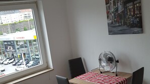Ein Hotelzimmer mit einem Bett, einem Tisch mit kariertem Tuch, zwei Stühlen, einem Ventilator und einem Fenster mit Blick auf eine Straße und geparkte Autos.