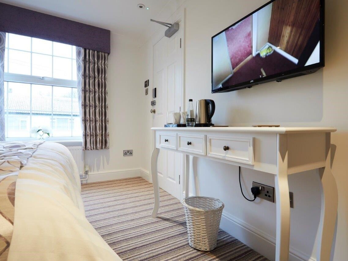 Habitación de hotel con cama, escritorio con cajones, televisor montado en la pared, ventana con cortinas y un cesto de basura.