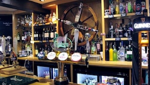 A bar area with a large wheel decoration, various liquor bottles, and branded taps. - The Rose & Crown Free House (Clacton-on-Sea)