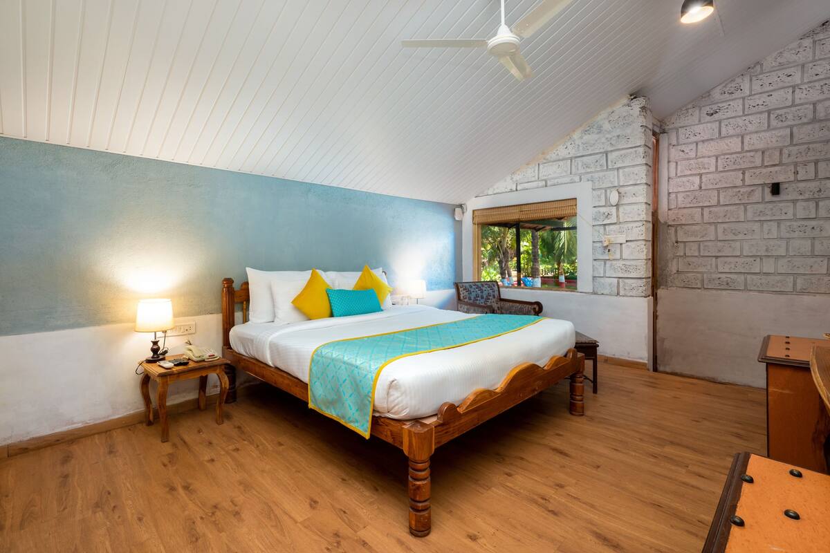Une chambre avec un lit en bois, une table de chevet avec une lampe, une fenêtre donnant sur des palmiers et un mur en briques.