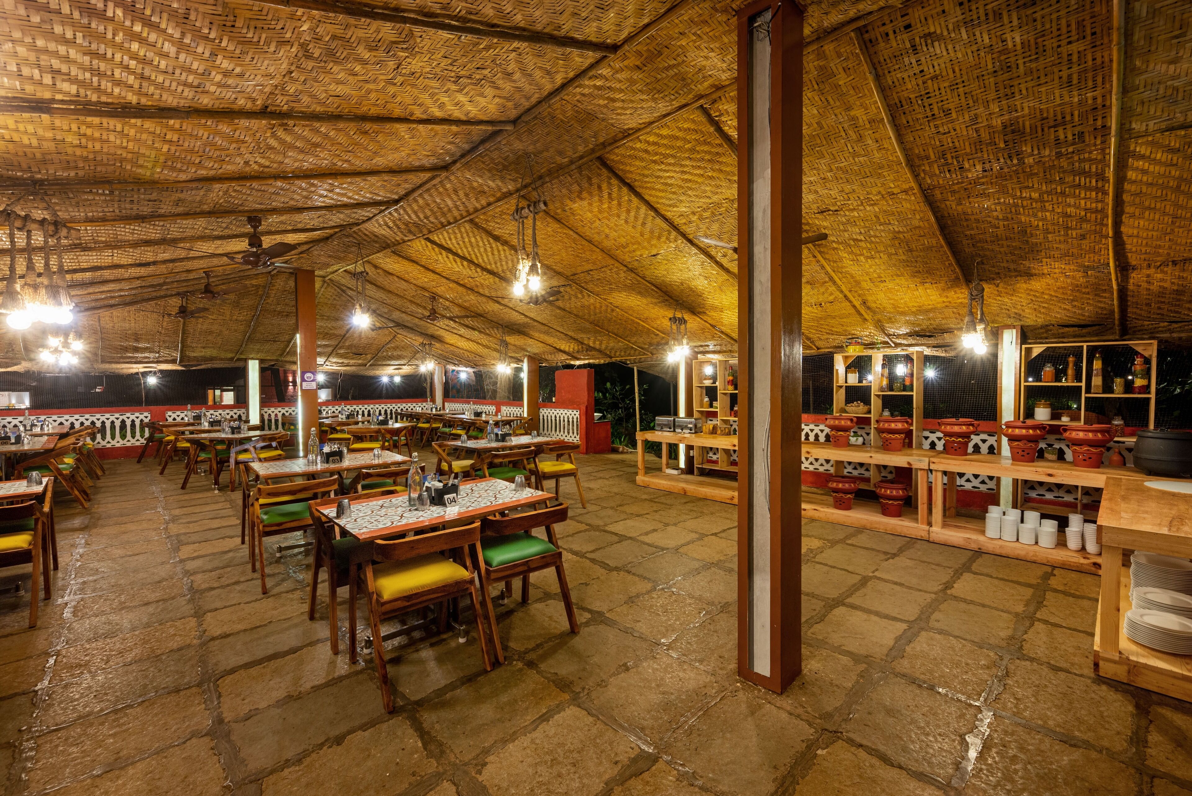 A spacious restaurant with high ceilings and a thatched roof, featuring multiple dining tables and chairs, and shelves with kitchenware.