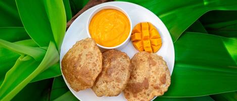 A plate with three fried items, a bowl of mango puree, and a mango on a white plate surrounded by green leaves.