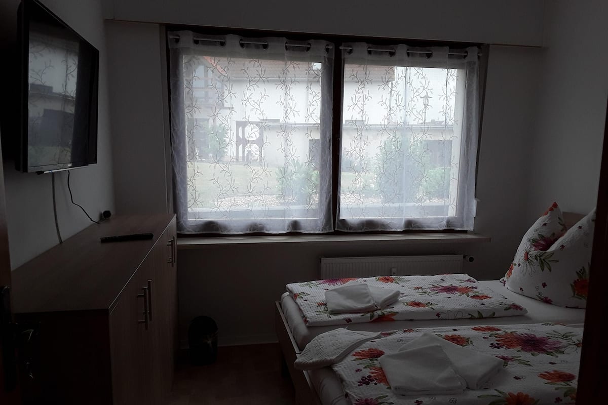 A bedroom with a bed, a window with curtains, a radiator, a TV mounted on the wall, and a small cabinet.