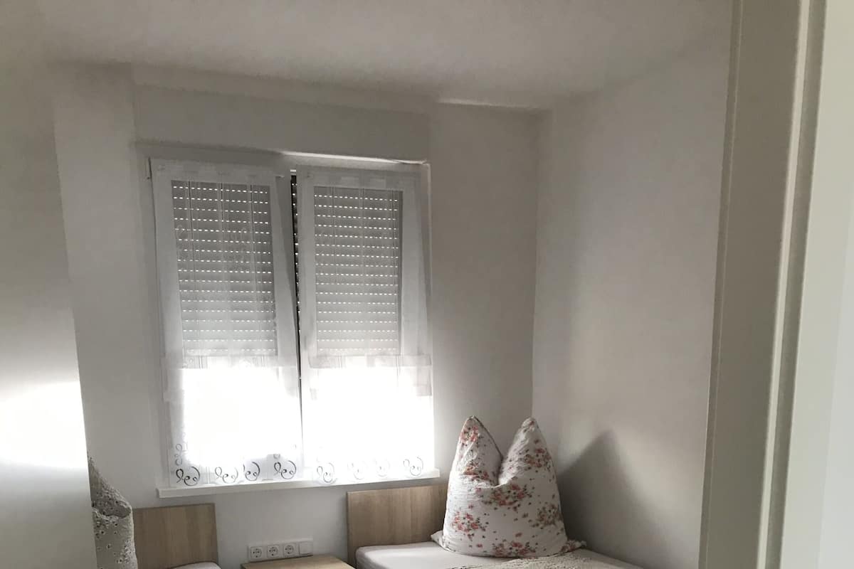 A small, compact bedroom with two single beds, a bedside table, a nightstand, and a window with blinds.
