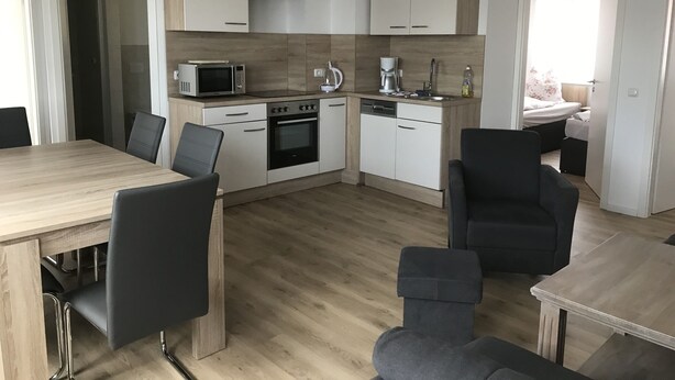 Una cocina moderna con armarios blancos, microondas, horno y cafetera. Hay una mesa de comedor con sillas y una pequeña zona de estar con sofá.