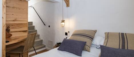 Un dormitorio con un escritorio de madera, una silla, una cama con almohadas, una lámpara de pared y una escalera.