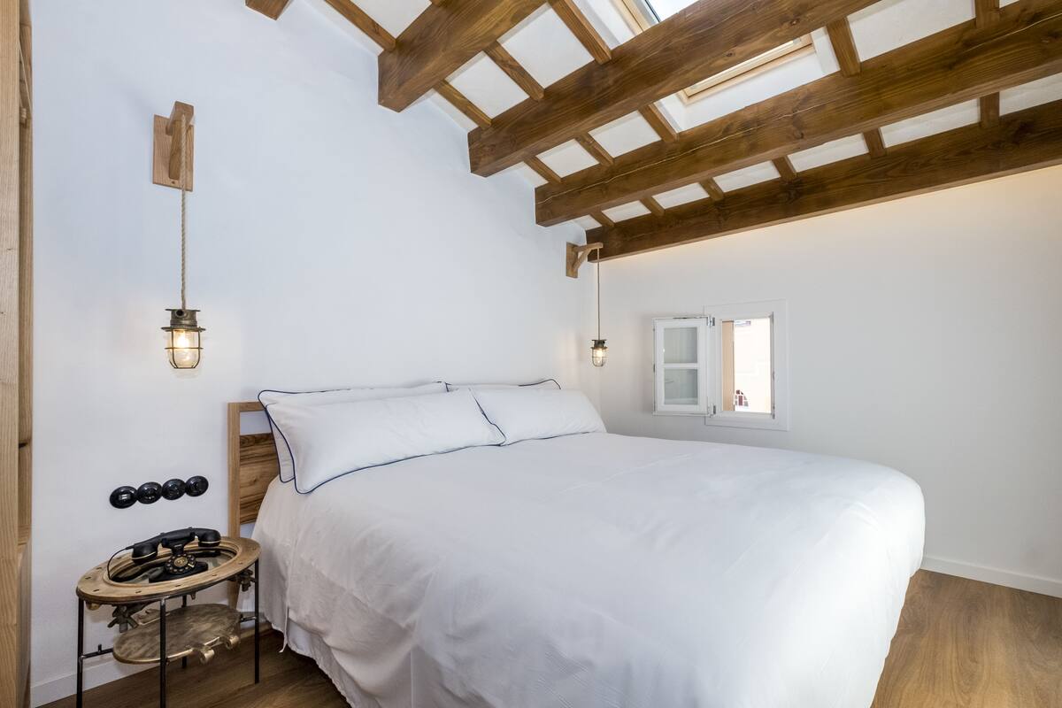 A bedroom with a wooden ceiling, a bed with white bedding, a small round table with a lamp, and a window with a view.