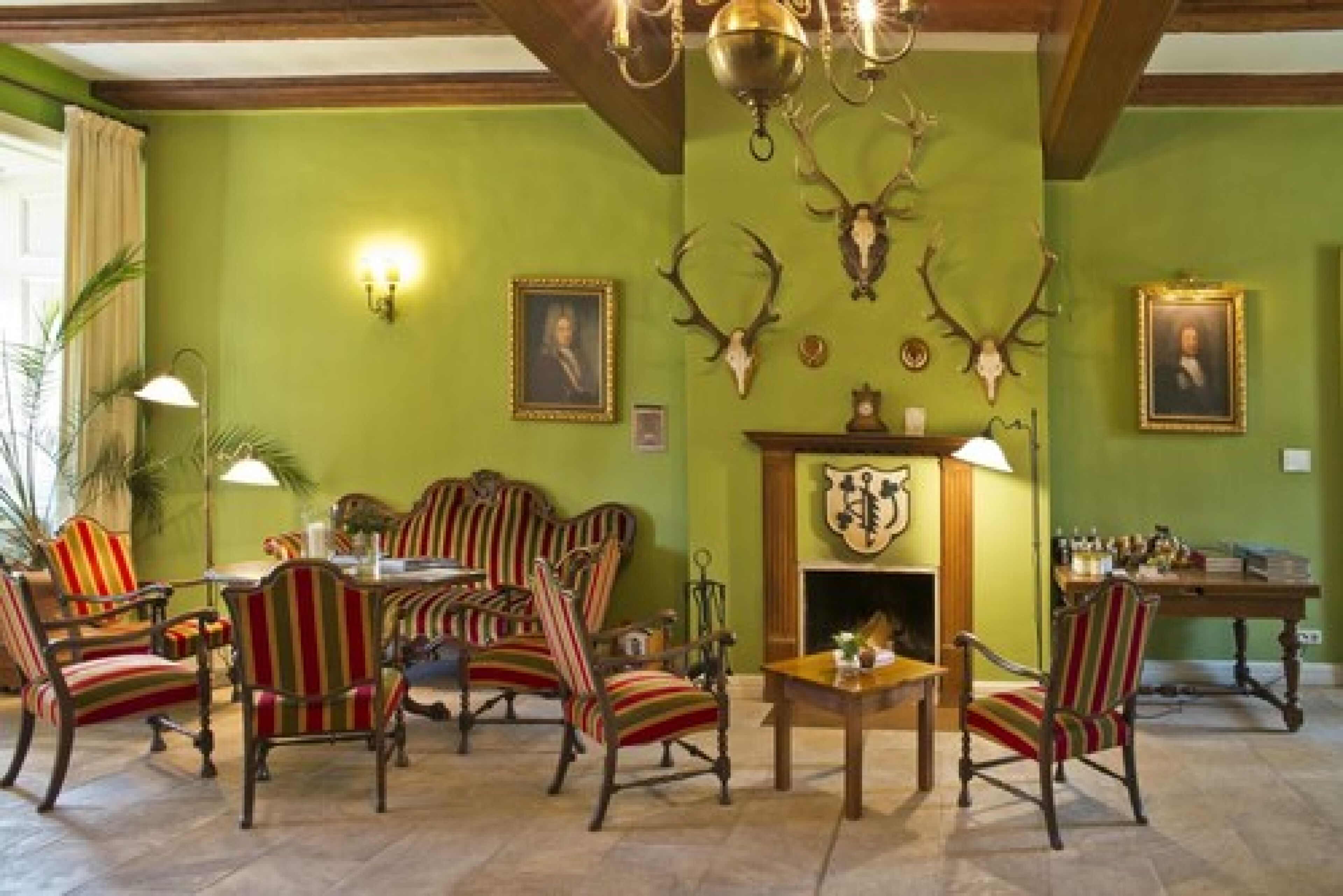 A room with green walls, striped chairs, a fireplace with a coat of arms, and mounted deer heads.