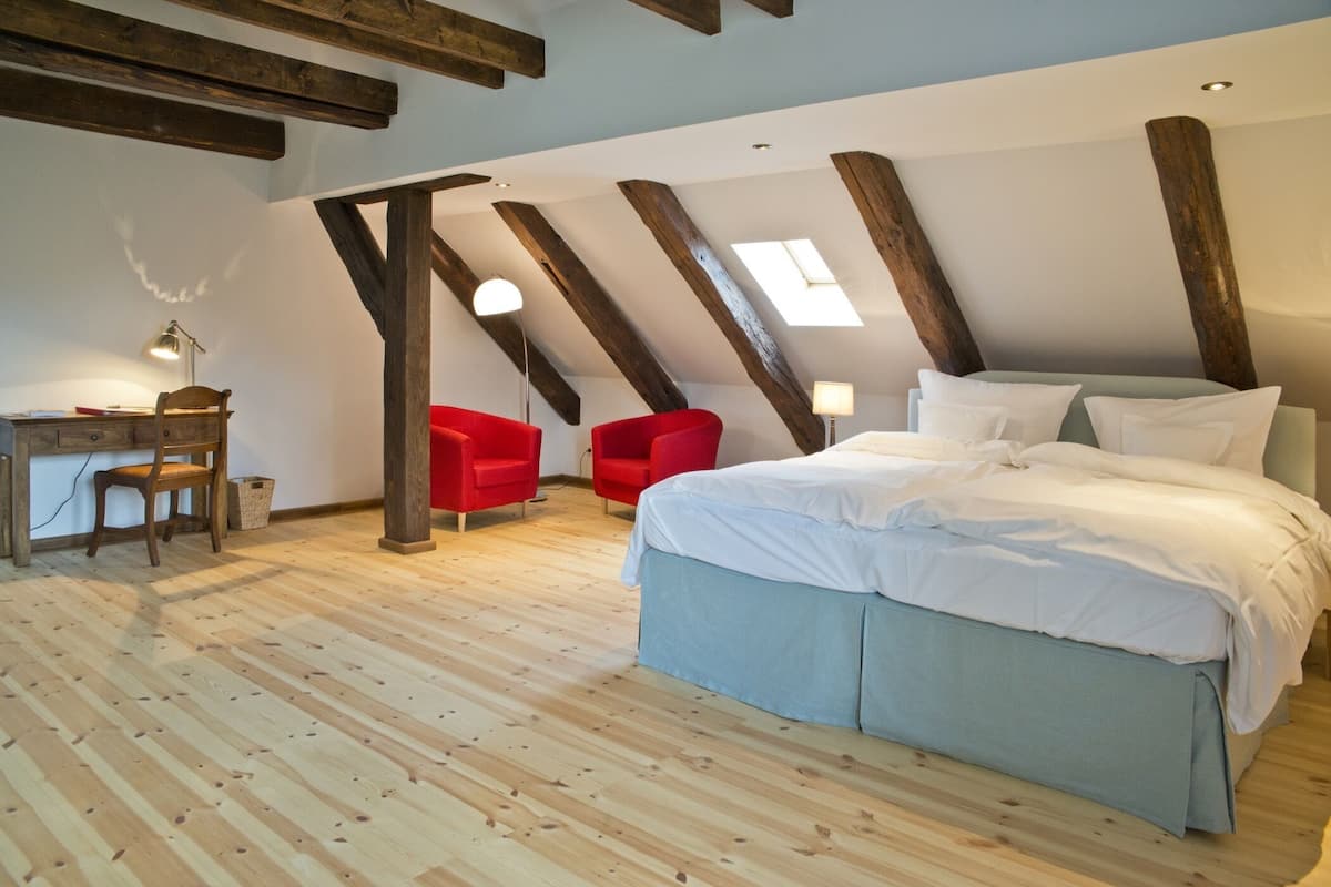 Un dormitorio con piso de madera, una cama con ropa de cama blanca, un escritorio con lámpara y sillas rojas.