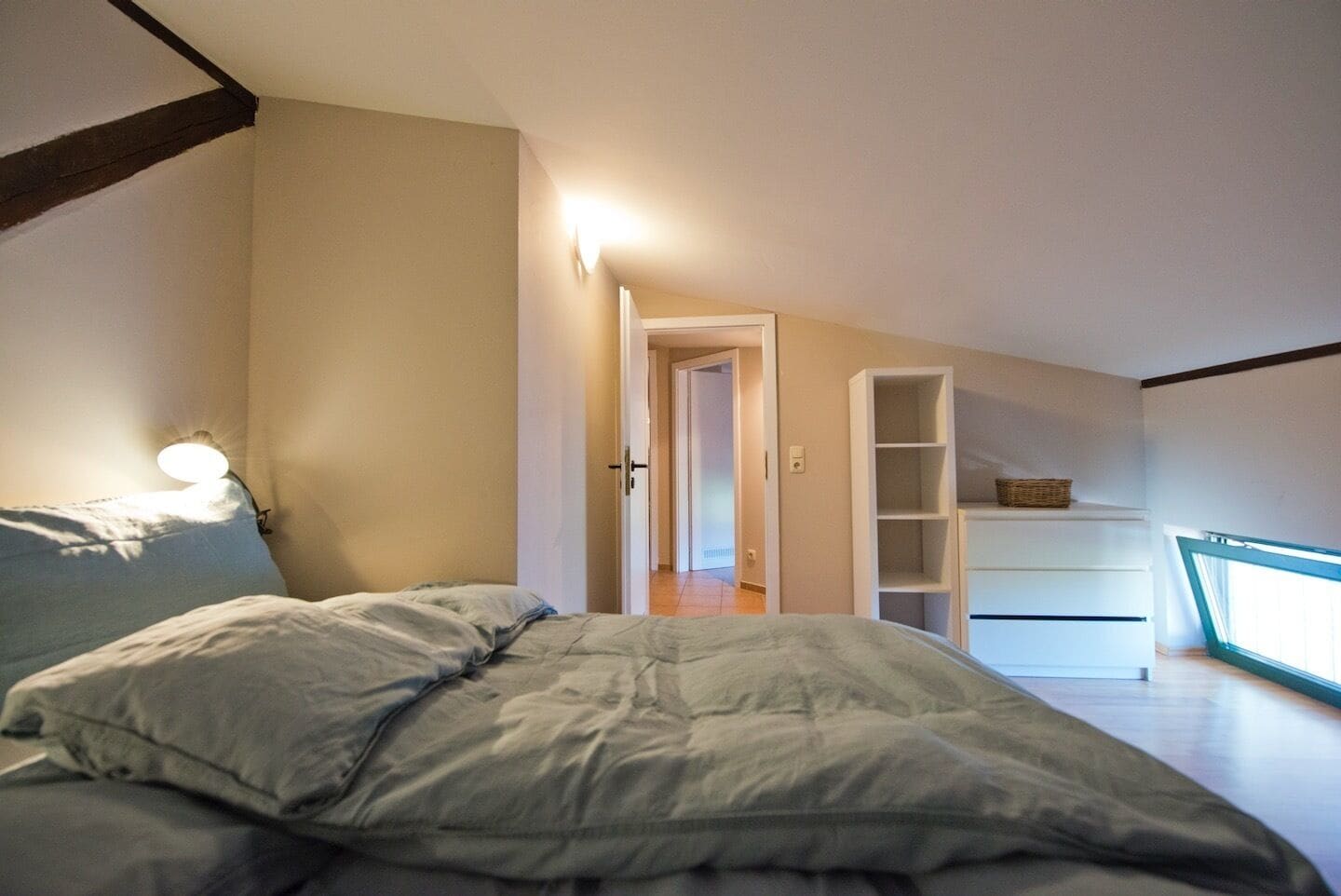 Un dormitorio con cama, mesita de noche, lámpara de pared, estantería blanca y puerta que da a otra habitación.