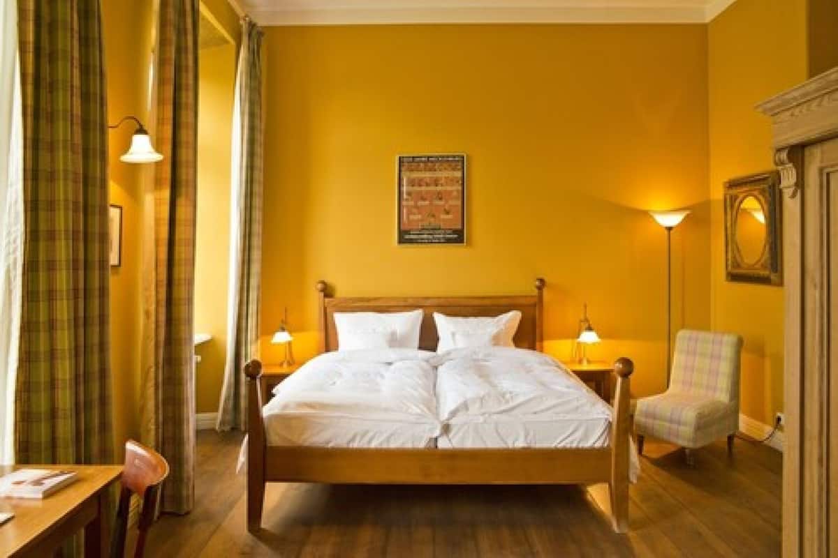 Una habitación de hotel con una cama de madera, dos lámparas de noche, un sillón a cuadros y un cartel en la pared.