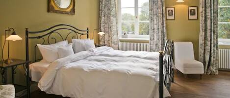 A neatly made bed in a room with a large window, a chair, and a small table with a lamp.