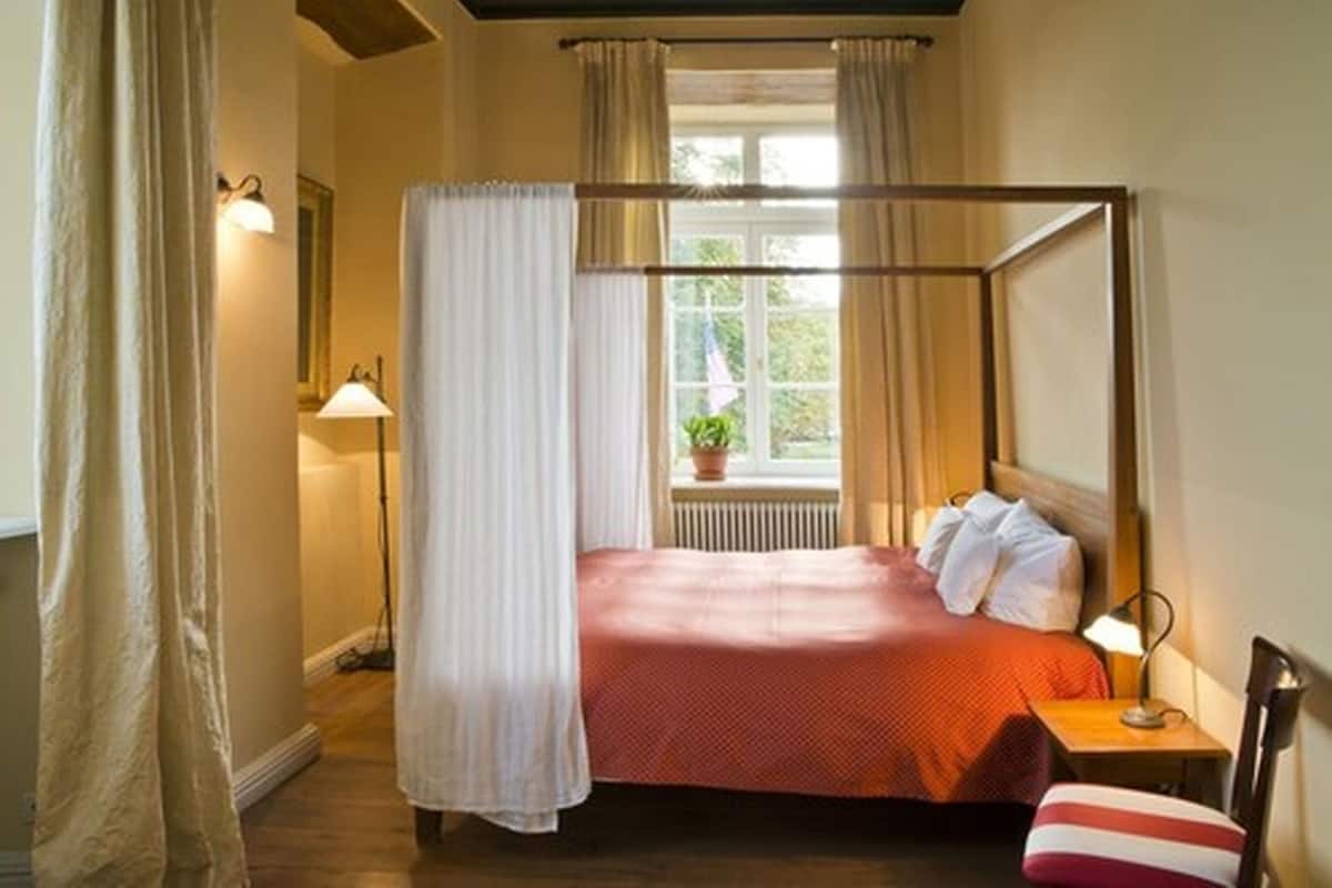 Una cama con dosel, con una colcha roja, una mesita de noche con lámpara y una ventana con cortinas.