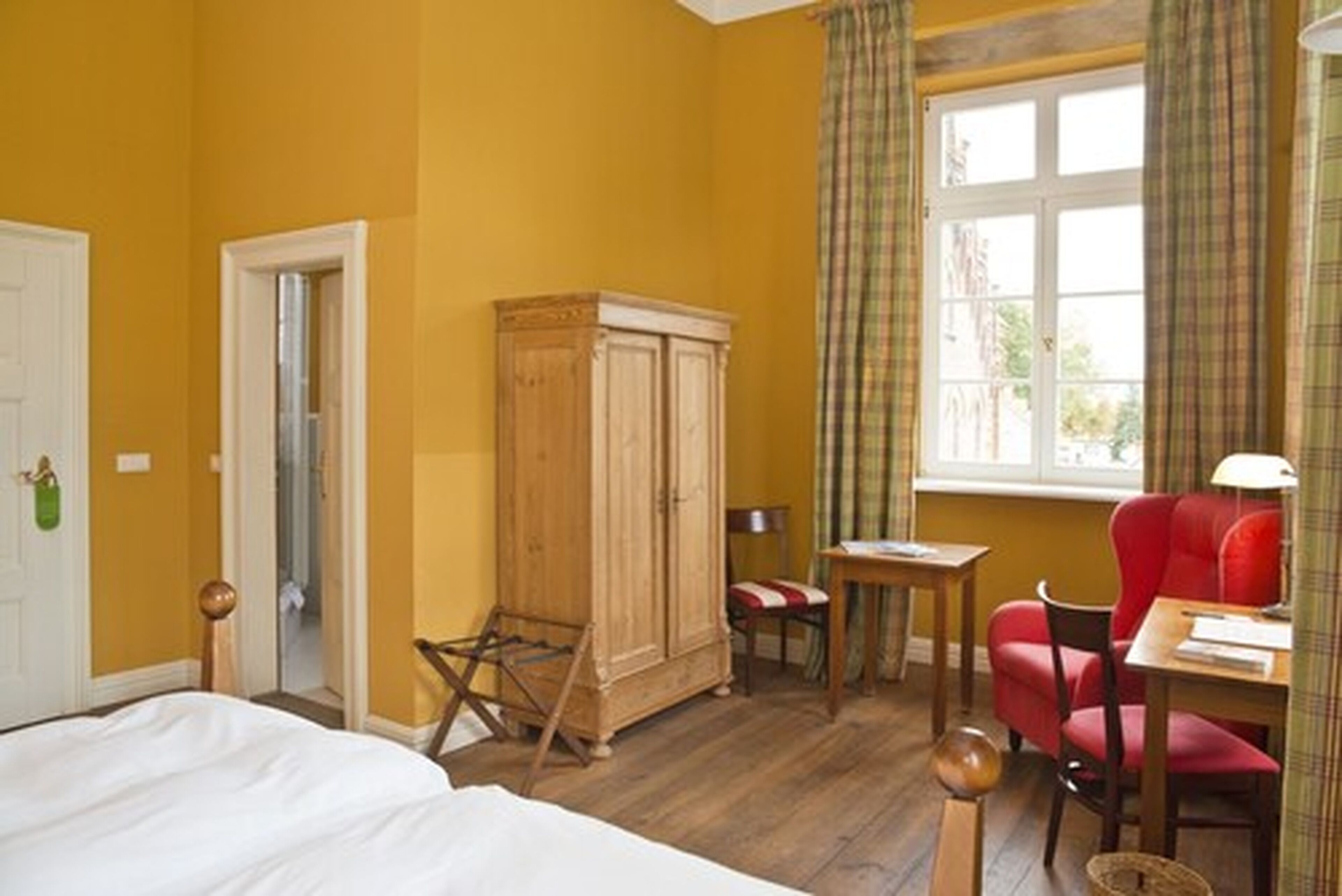 A room with a bed, a wooden wardrobe, a small table with a lamp, a red chair, and a window with curtains.