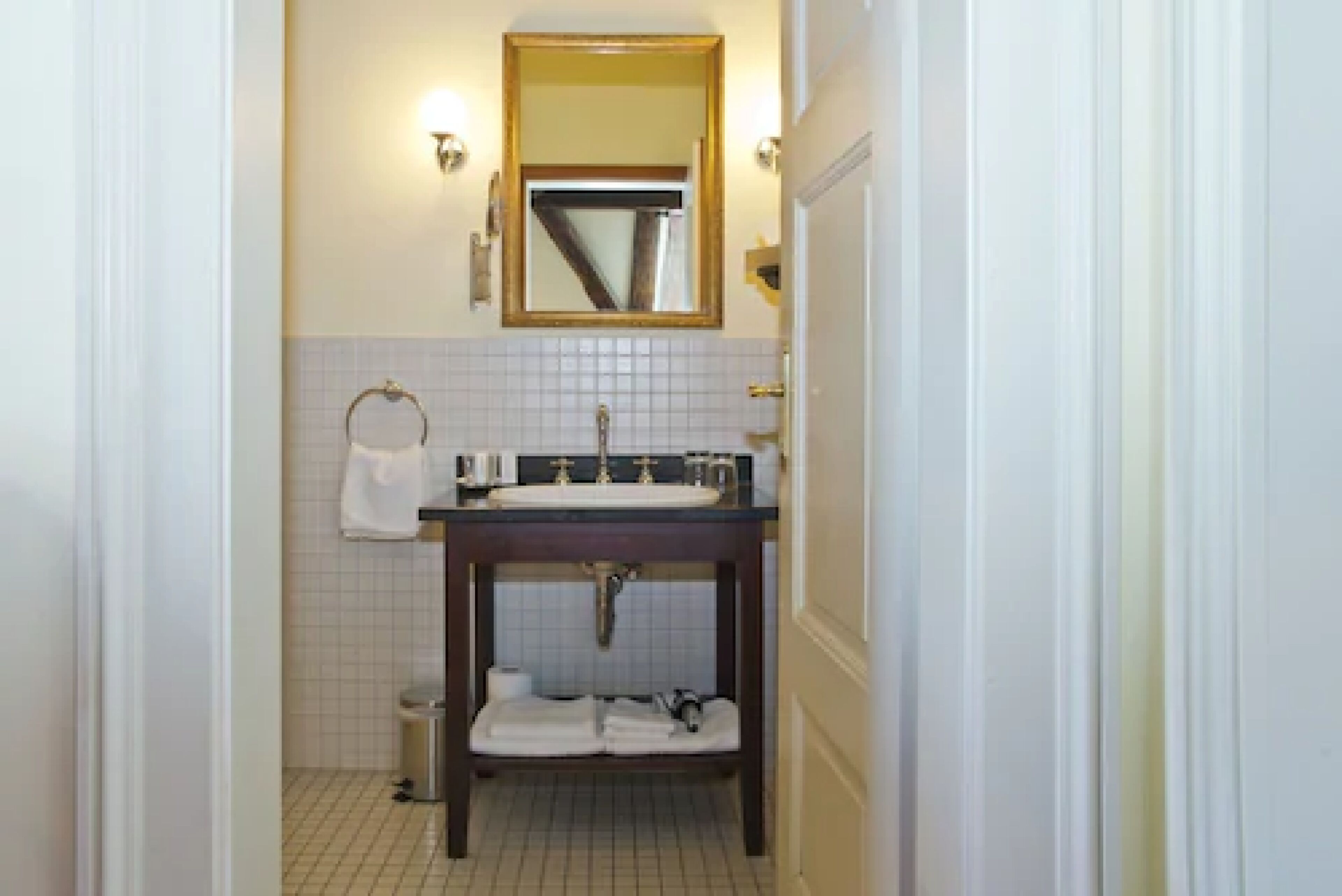 Un baño con tocador de madera oscura, paredes de azulejos blancos, un espejo con marco dorado y una toalla blanca colgada en la pared.