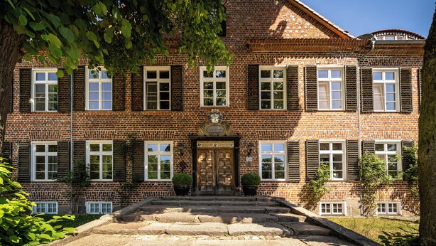 Ein großes Backsteingebäude mit symmetrischer Fassade, zahlreichen Fenstern mit Fensterläden und einem zentralen Eingang mit Stufen.