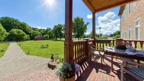 Eine überdachte Terrasse mit Backsteinboden, Holzmöbeln und Blick auf eine grüne Rasenfläche sowie ein Backsteingebäude im Hintergrund.
