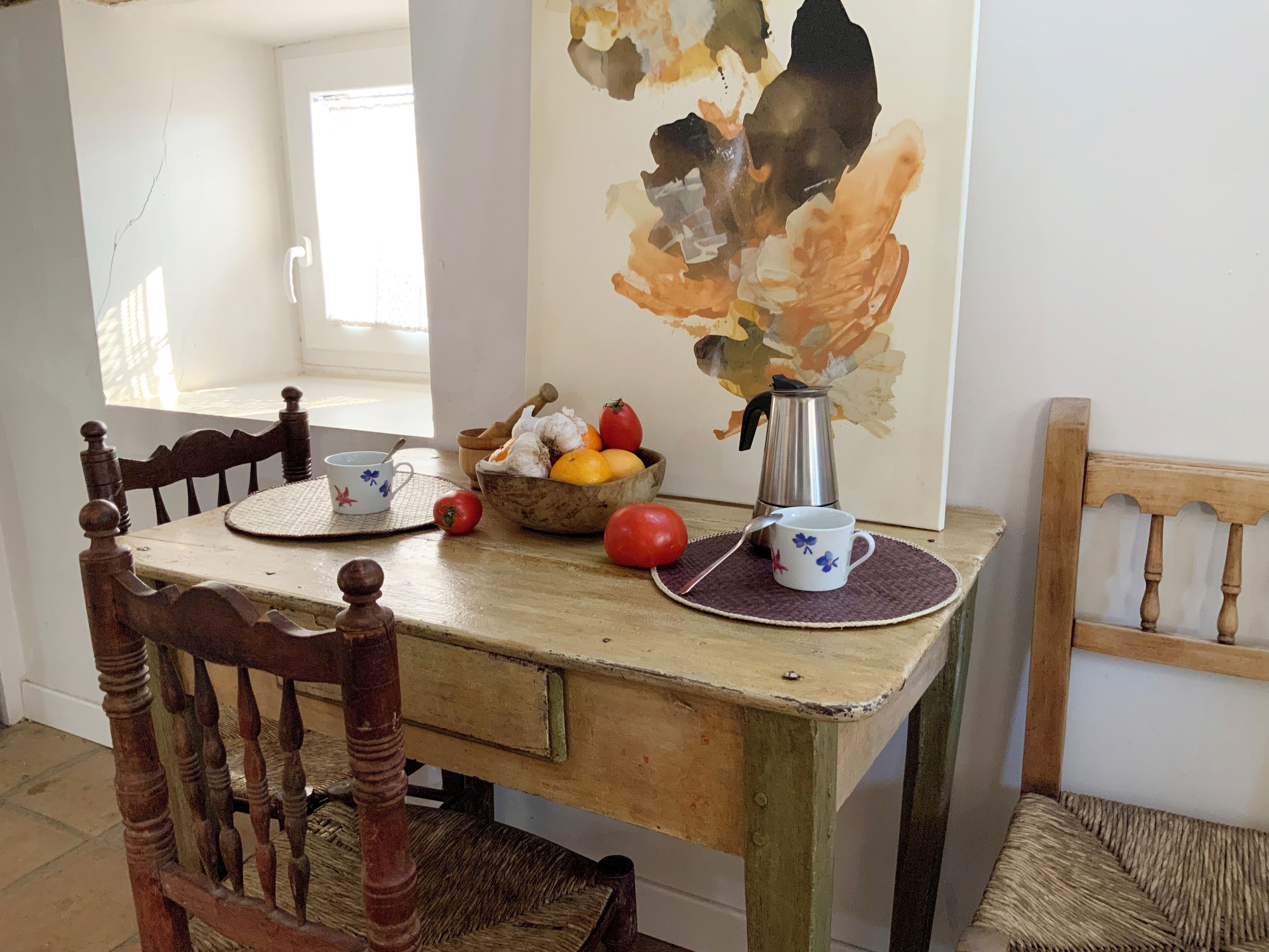 Una mesa de comedor de madera con acabado rústico, un cuenco de fruta, una cafetera y una taza sobre un posavasos tejido.