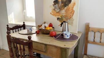 Una mesa de comedor de madera con acabado rústico, un cuenco de fruta, una cafetera y una taza sobre un posavasos tejido.