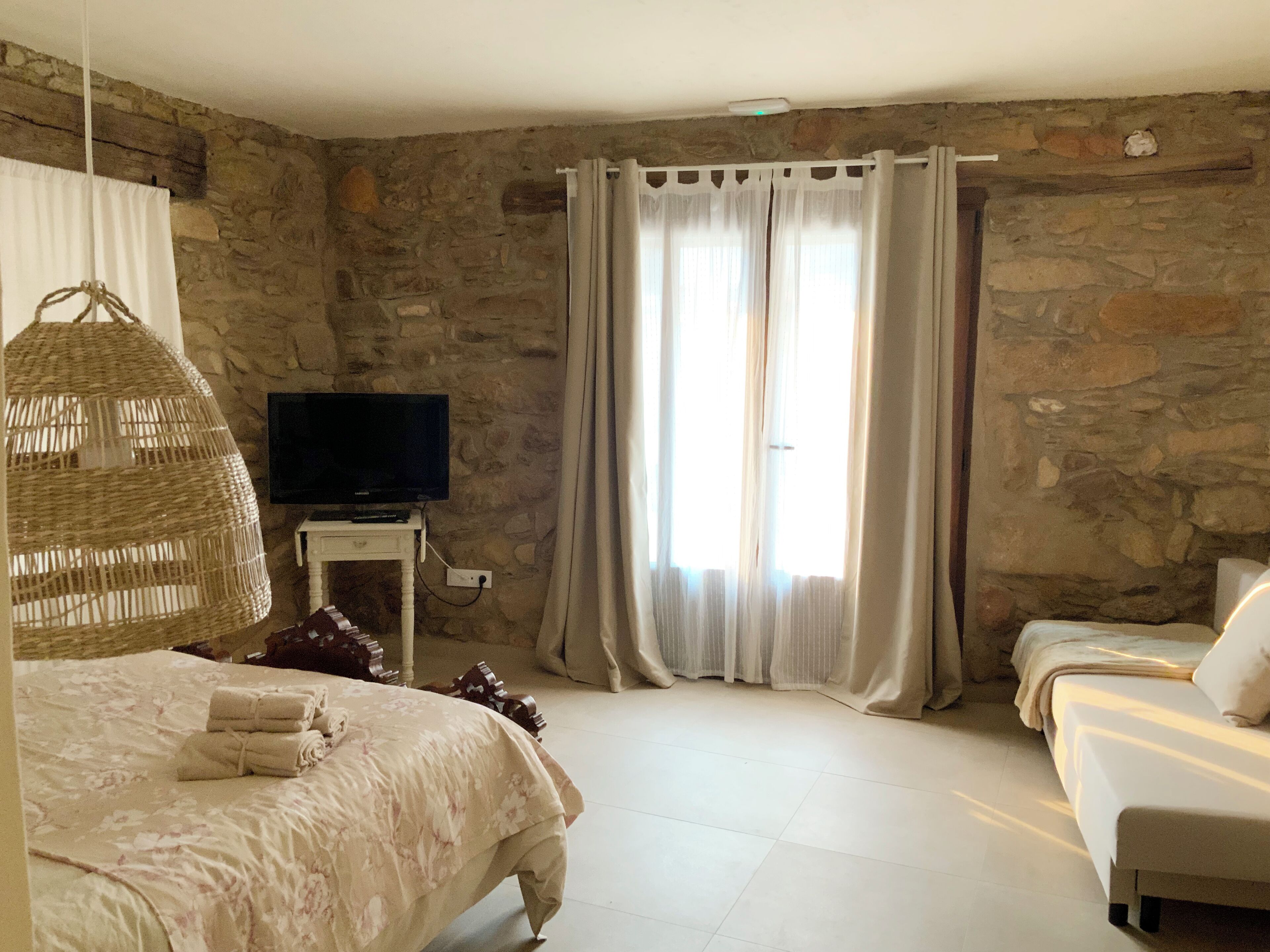 Un dormitorio con pared de piedra, un televisor sobre una mesita, una cama con cubrecama floral y un sofá con funda blanca.