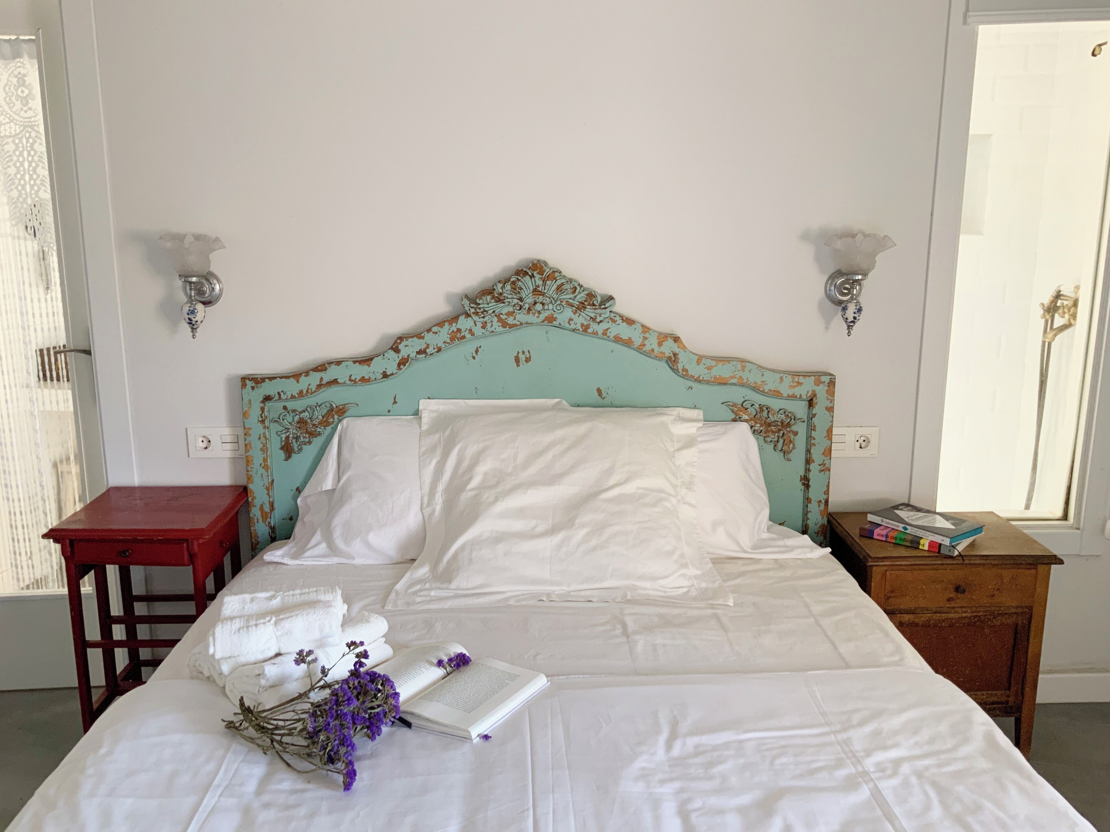 A neatly made bed with white linens, a book, and purple flowers on a bedside table.