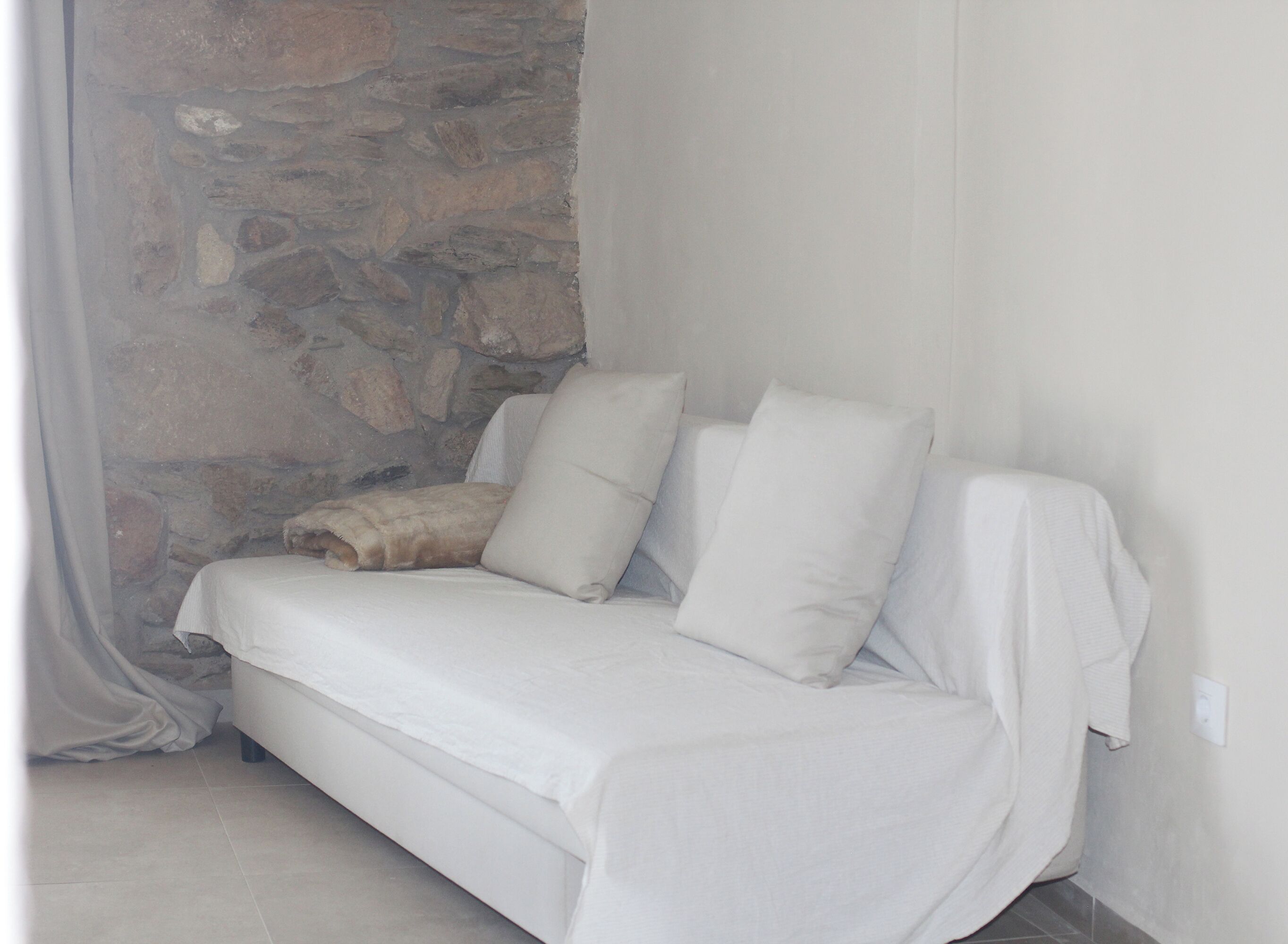 A white sofa with cushions against a stone wall and a white curtain.