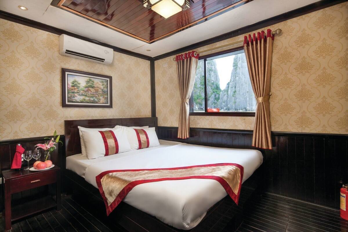 A hotel room with a large bed, a nightstand with a vase and a red cloth, a painting on the wall, and a window with curtains.