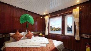 Une chambre d’hôtel avec un grand lit, un éventail décoratif vert, deux fenêtres avec des rideaux et une vue sur les montagnes.