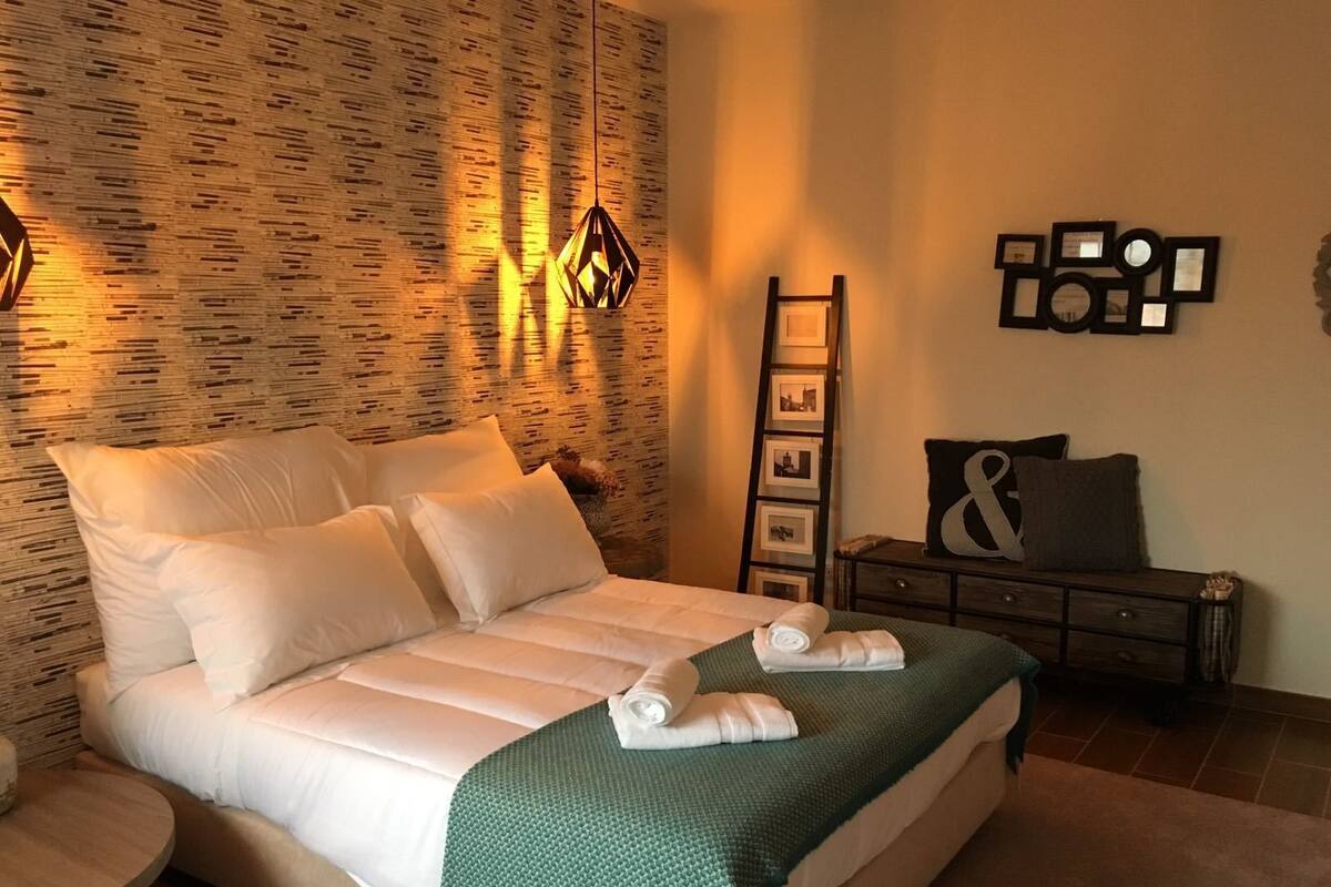Cama bem arrumada com lençóis brancos e manta verde, parede de madeira com padrão texturizado e escada fixada na parede com quadros.