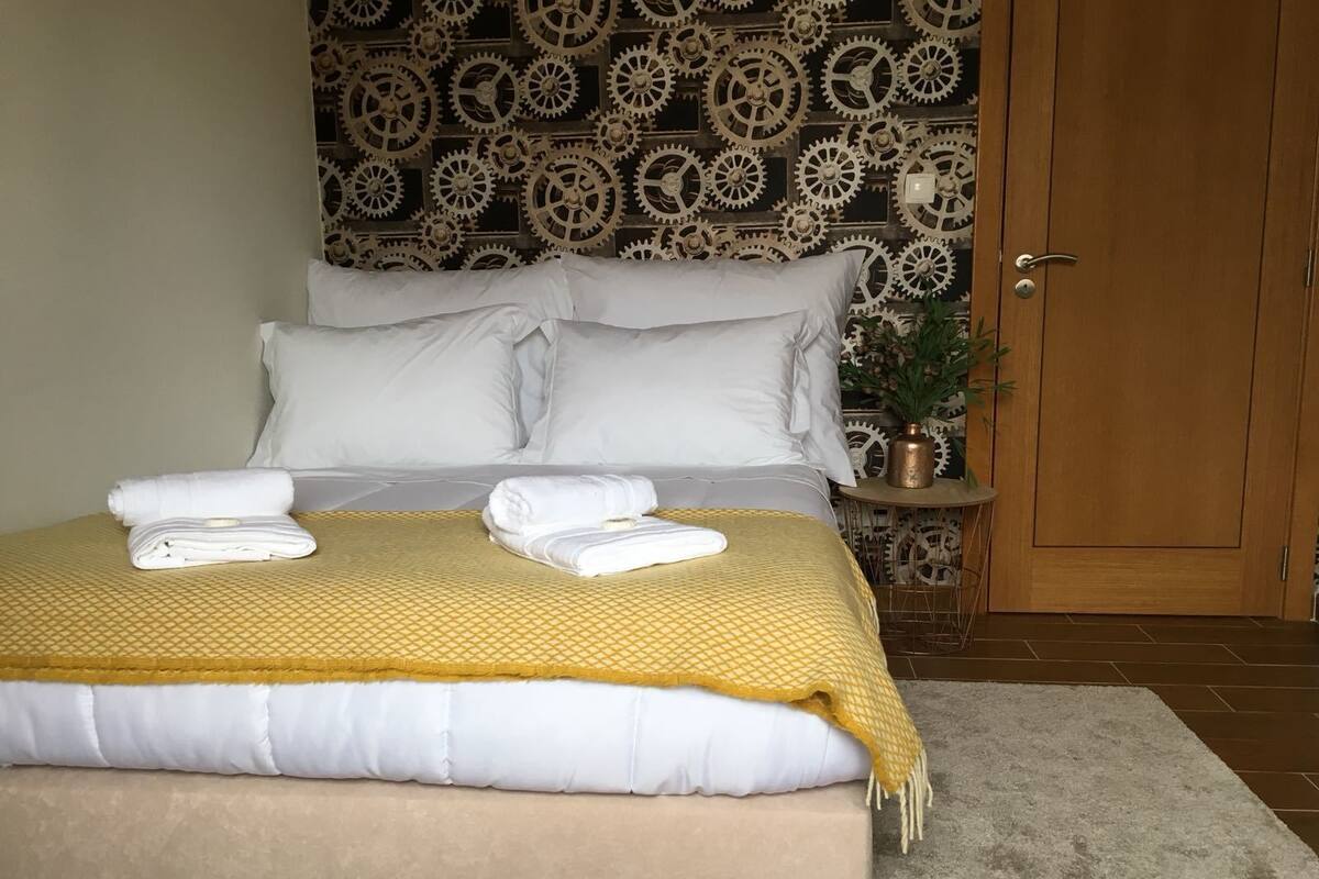 Cama bem arrumada com lençóis brancos e um cobertor amarelo, uma porta de madeira e uma parede com padrão de engrenagens.