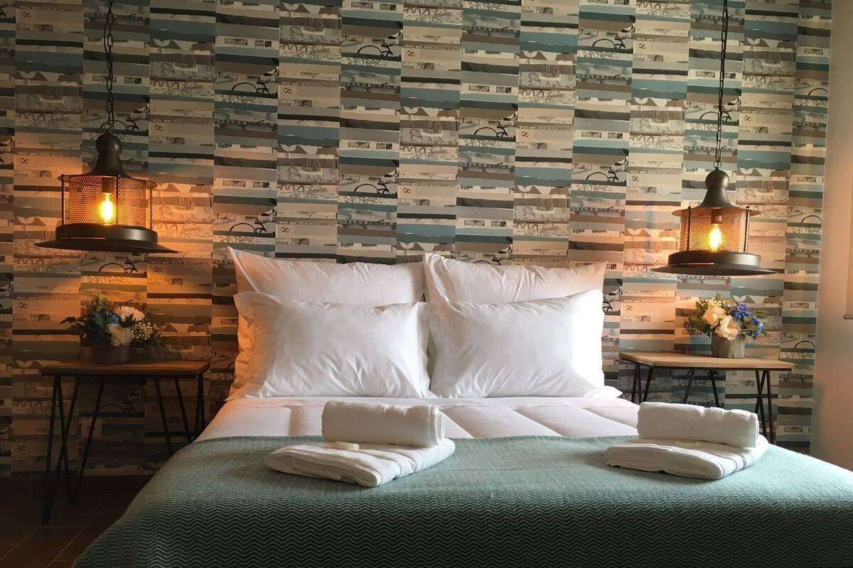 Cama bem arrumada com lençóis brancos e manta verde, com duas mesinhas pequenas com arranjos florais ao lado.